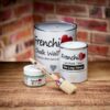 Frenchic Oval Brush with red ferrule shown alongside Frenchic Chalk Wall Paint, Lazy Range Furniture Paint, and Al Fresco paint tins on wooden surface with brick background