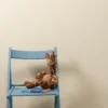A plush giraffe toy sitting on a blue wooden chair against a plain beige wall, with a children's book underneath.