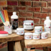 Collection of Frenchic products including Frensheen metallic powders, furniture wax, Finishing Coat, and application tools, displayed on wooden stools against a red brick wall.