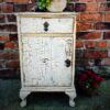 Antique wooden cabinet with distressed, cracked white paint finish against a brick wall, decorated with a flower pot on top and a potted plant beside.
