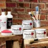 Frenchic finishing products including Tuff Top Coat, Finishing Coat, Furniture Wax tins, brushes, and sponge displayed on rustic wooden stools against a brick wall background