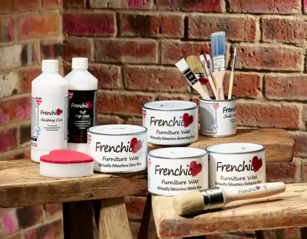 Frenchic finishing products including Tuff Top Coat, Finishing Coat, Furniture Wax tins, brushes, and sponge displayed on rustic wooden stools against a brick wall background
