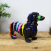 Colorful striped dachshund figurine with a green nose on a wooden surface, next to a potted plant and a decorative light.