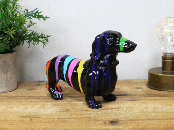 Colorful striped dachshund figurine with a green nose on a wooden surface, next to a potted plant and a decorative light.