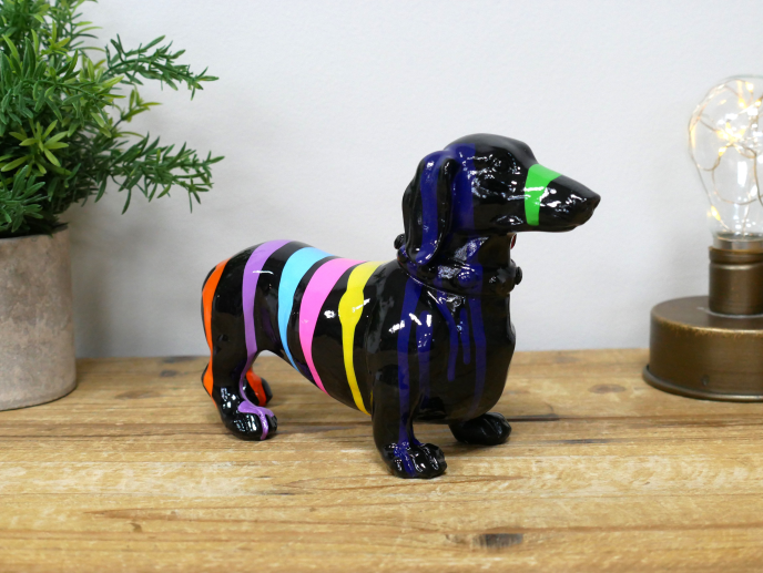 Colorful striped dachshund figurine with a green nose on a wooden surface, next to a potted plant and a decorative light.