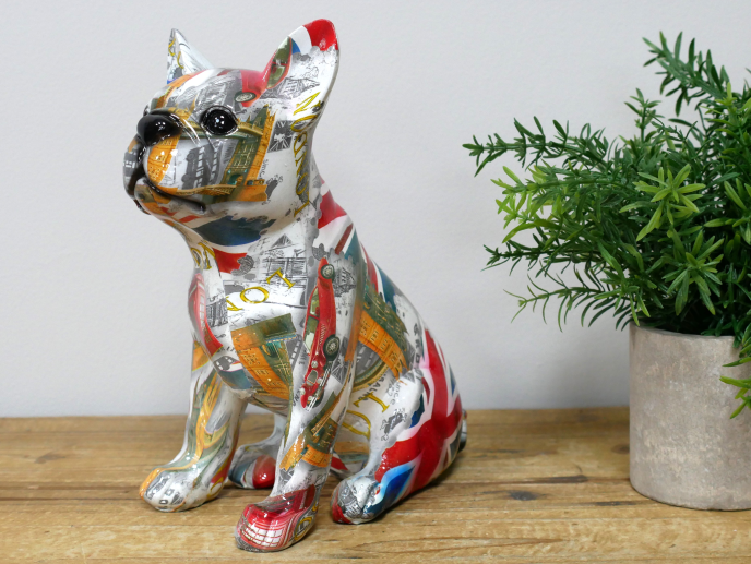 Decorative dog figurine with a collage of UK and American flags and cityscape images, placed on a wooden surface next to a green potted plant.