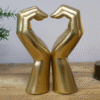 Decorative gold hand sculptures forming a heart shape on a wooden surface with a small plant and a light bulb in the background.