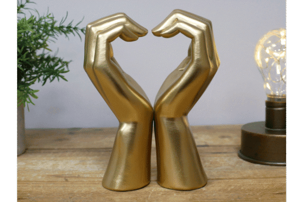 Decorative gold hand sculptures forming a heart shape on a wooden surface with a small plant and a light bulb in the background.
