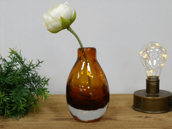 Small amber glass vase with a white flower, placed on a wooden surface with decorative greenery and a vintage-style light bulb.