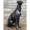 Decorative black sitting whippet sculpture made of metal or resin, placed on a wooden floor against a brick wall background.
