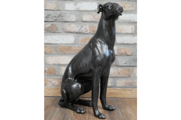 Decorative black sitting whippet sculpture made of metal or resin, placed on a wooden floor against a brick wall background.