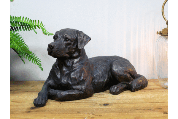 Decorative black dog sculpture lying on a wooden surface with a green fern plant nearby.