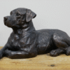Lying dog sculpture made of dark material, resting on a wooden surface against a plain wall.