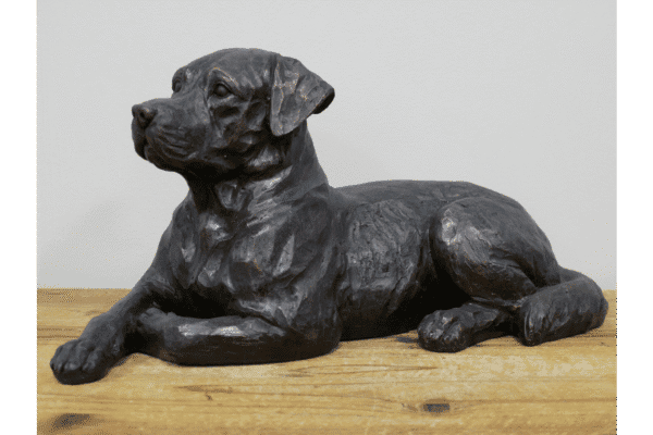 Lying dog sculpture made of dark material, resting on a wooden surface against a plain wall.