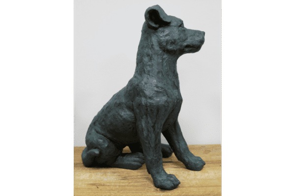 Bronze sculpture of a sitting dog with detailed features, placed on a wooden surface against a plain white background.