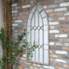 Arch rustic mirror with metal frame hanging on a brick wall, surrounded by green plants.