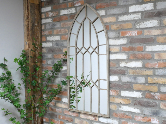 Arch rustic mirror with metal frame hanging on a brick wall, surrounded by green plants.