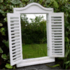 White shutter mirror with a decorative top, mounted on a garden wall, reflecting greenery and trees.