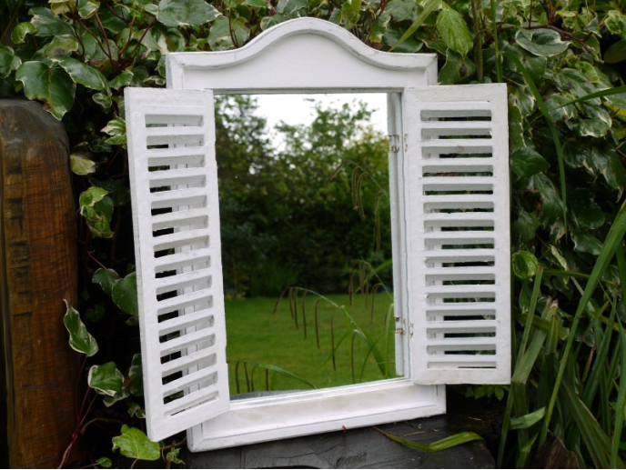White shutter mirror with a decorative top, mounted on a garden wall, reflecting greenery and trees.