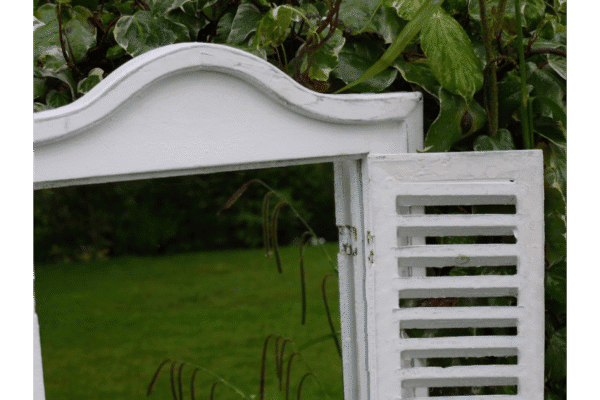 White shutter mirror with decorative top, set against green foliage, showcasing home decor and mirror design.