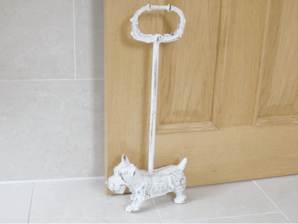 Decorative metal door stop shaped like a Westie dog with a handle, placed on a tiled floor near a wooden door.