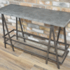 Industrial console table with a metal frame and a concrete top, featuring a lower wire mesh shelf, set against a brick wall background.