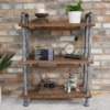 Industrial pipe shelf unit with three wooden shelves against a brick wall, decorated with plants, books, a clock, and a dog figurine.