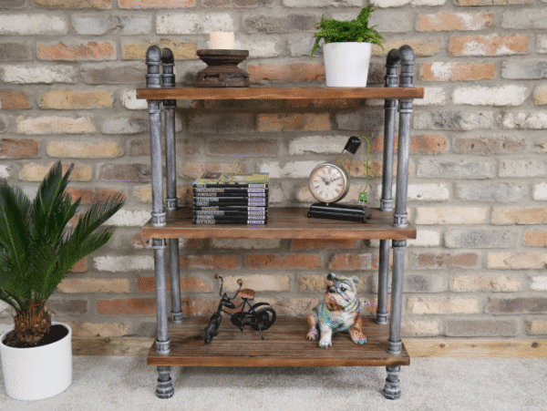 Industrial pipe shelf unit with three wooden shelves against a brick wall, decorated with plants, books, a clock, and a dog figurine.
