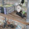 Industrial pipe shelf unit displaying magazines, a vintage clock, and decorative items against a brick wall.