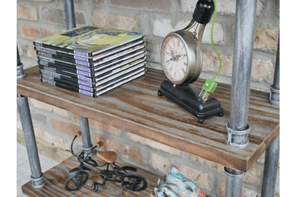 Industrial pipe shelf unit displaying magazines, a vintage clock, and decorative items against a brick wall.