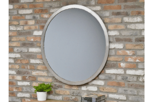 Industrial round silver mirror with a concrete frame hanging on a brick wall, with a small potted plant and books below.