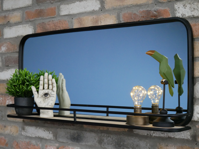 Decorative wall mirror with black frame, featuring potted plant, hand sculpture, and modern light bulbs on a black shelf against a brick wall.