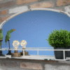 Aged white mirror with a shelf, decorated with green bird figurines, two small lamps, and a potted green plant, mounted on a brick wall.