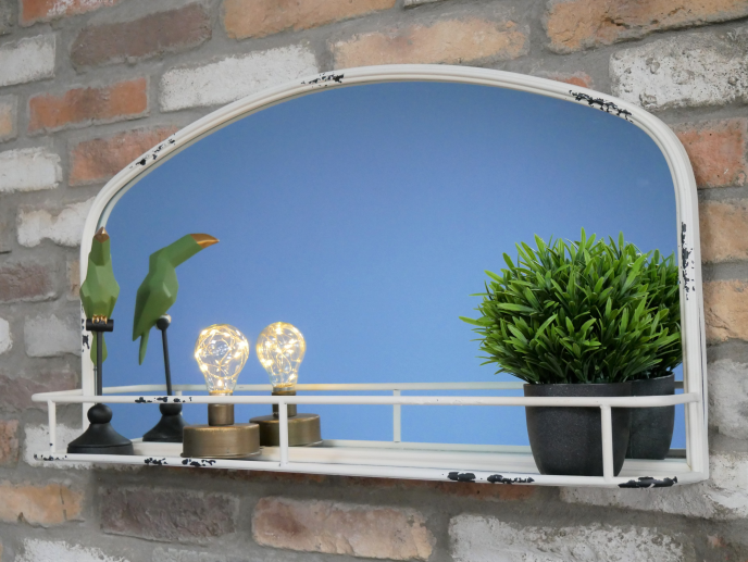 Aged white mirror with a shelf, decorated with green bird figurines, two small lamps, and a potted green plant, mounted on a brick wall.
