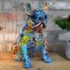 Colourful graffiti-style bulldog sculpture with vibrant patterns and bold colours, displayed on a wooden surface with a brick wall background.