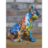 Colourful graffiti-style sitting bulldog sculpture on wooden surface against brick wall background.