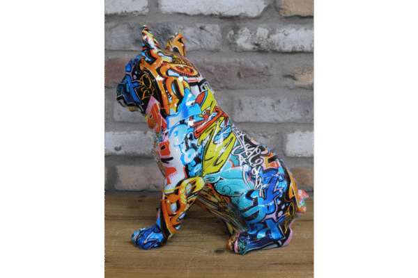 Colourful graffiti-style bulldog sculpture with vibrant patterns and bold colours, sitting on a wooden surface against a brick wall.