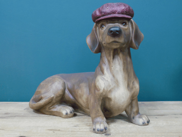 Decorative ceramic dog sculpture wearing a maroon cap, lying on a wooden surface against a blue background.