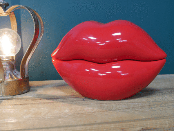 Red lips-shaped trinket box with glossy finish on wooden surface, decorative lamp in background.