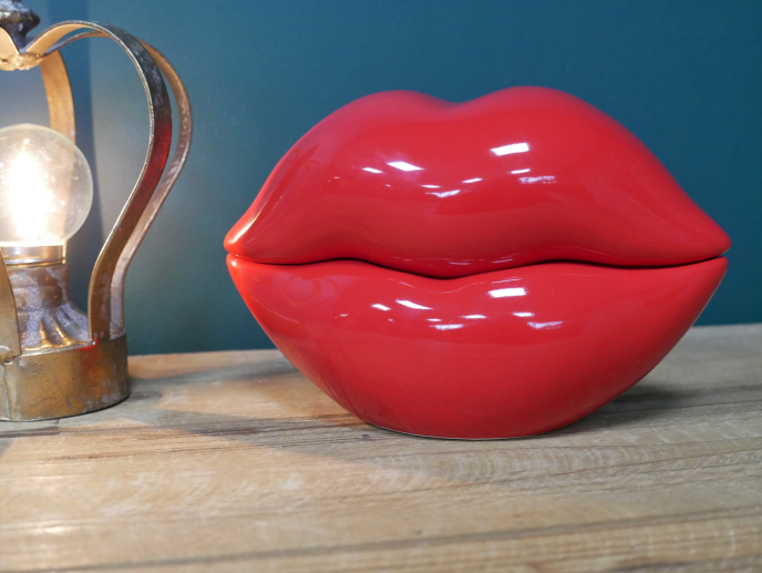 Red lips-shaped trinket box with glossy finish on wooden surface, decorative lamp in background.