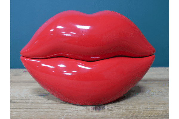 Red lips-shaped trinket box with glossy finish on a wooden surface against a blue background.