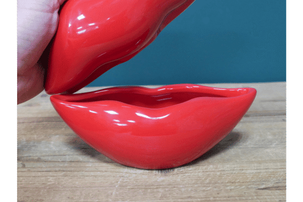 Red lips-shaped trinket box with a removable lid, glossy finish, on a wooden surface.