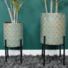 Set of two geometric patterned metal planters with black stands, containing tall green plants, placed against a teal wall on a grey shaggy rug.
