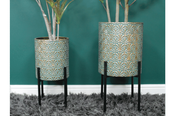 Set of two geometric patterned metal planters with black stands, containing tall green plants, placed against a teal wall on a grey shaggy rug.
