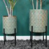 Two geometric metal planters with black stands, decorated with a repeating fan pattern, placed against a green wall on a grey shaggy rug.
