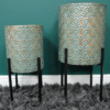 Pair of geometric metal planters with intricate fan pattern, small and large sizes, on black stands against a teal wall and grey shaggy rug.
