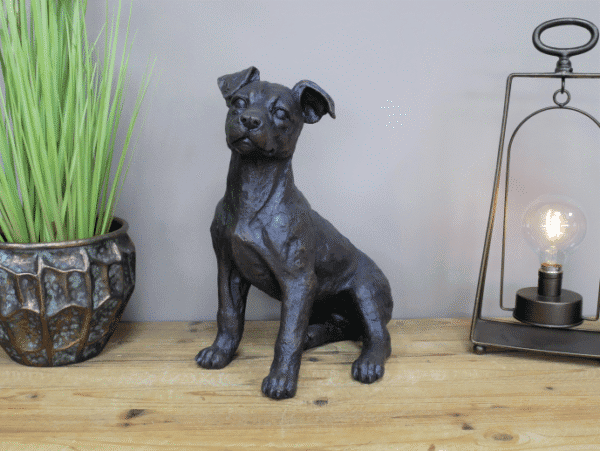 Gunmetal metallic sitting dog sculpture on wooden surface with potted plant and modern lamp in background.