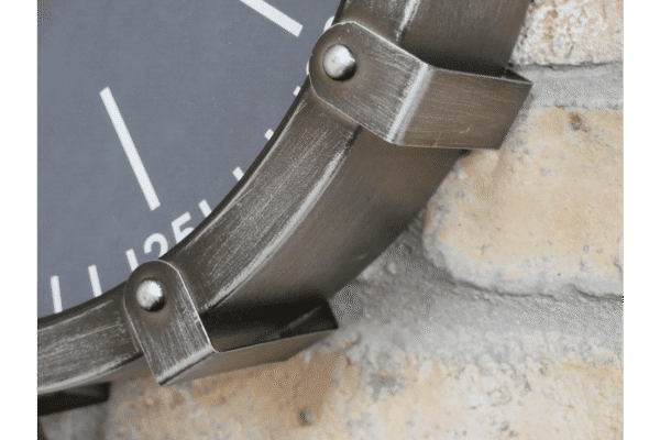 Close-up of an industrial-style cog clock with metallic finish and rivet details, mounted on a textured brick wall.