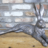 Metallic laying rabbit sculpture with a shiny silver finish, positioned on a wooden surface against a brick wall background.