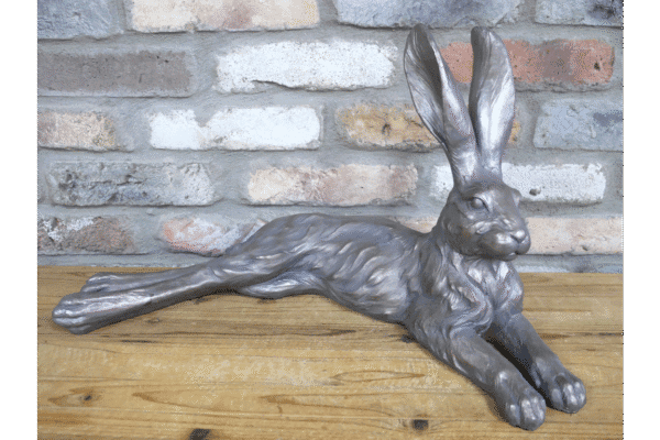 Metallic laying rabbit sculpture with a shiny silver finish, positioned on a wooden surface against a brick wall background.